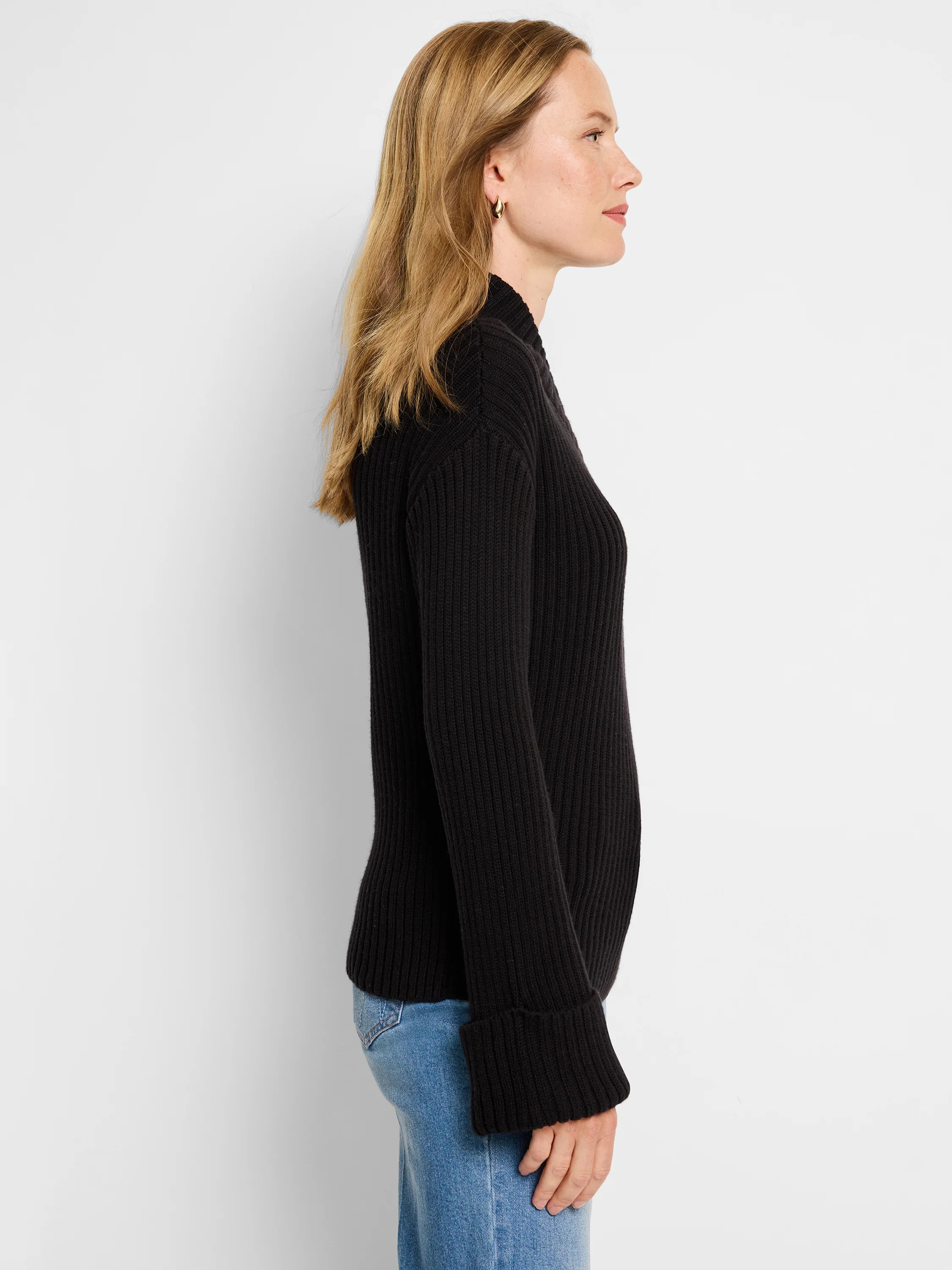 Black ribbed mock neck sweater. Long sleeves with folded cuffs. Side profile view.