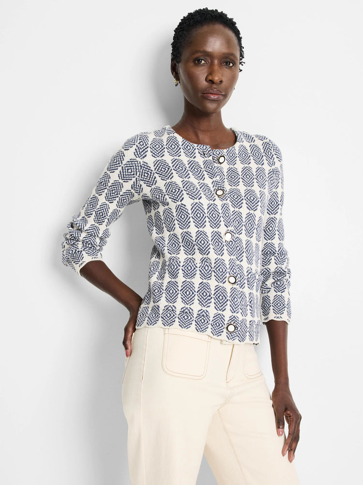 Off-white knit jacket with a navy blue geometric pattern and gold buttons. Long sleeves, close-fitting.