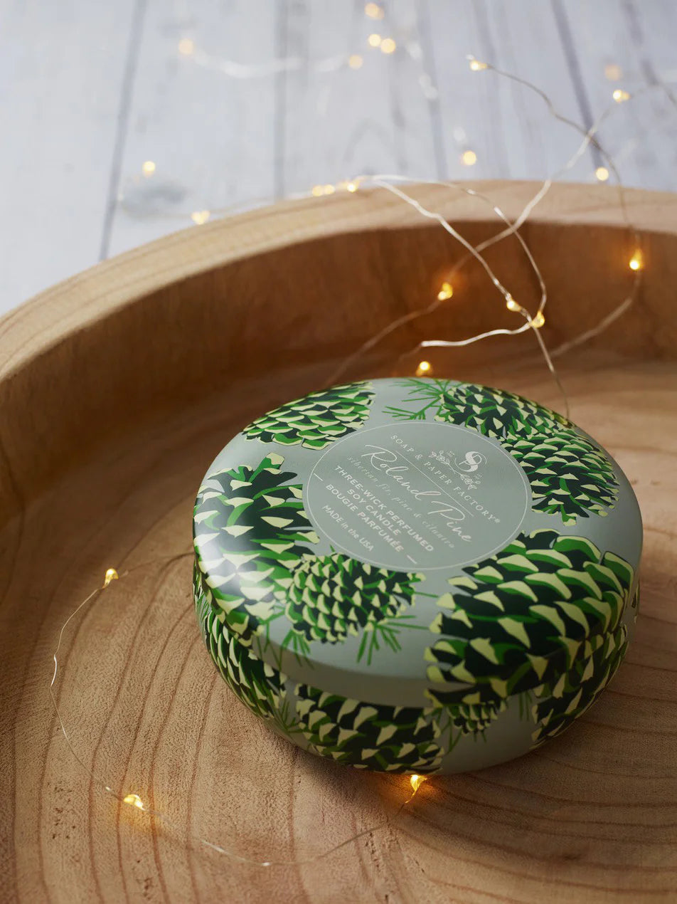 Round pale green candle tin with dark and light green pinecone pattern, featuring a white label. In a wooden bowl with...
