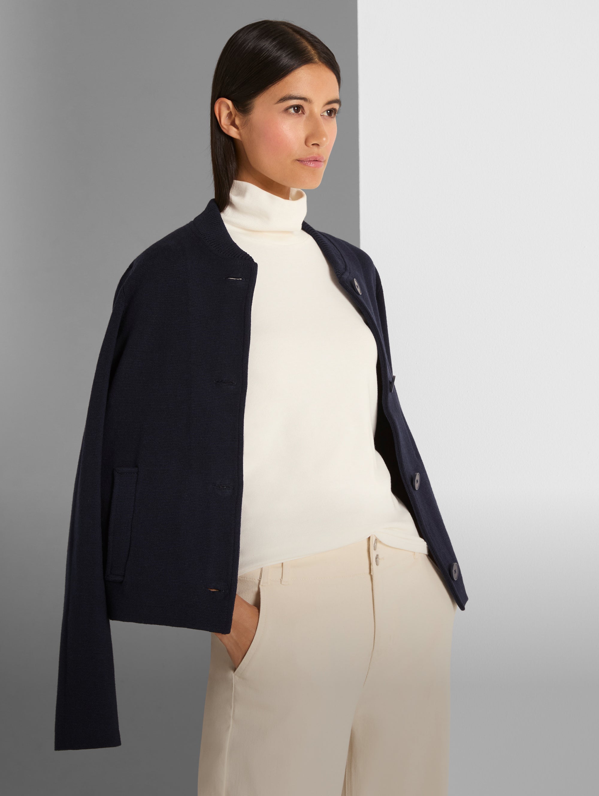 Woman wearing a navy blazer over a white turtleneck and beige pants on a gray background