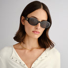 Model wears black oval sunglasses with thick frames and a gold logo paired with a slim fit white ribbed top.