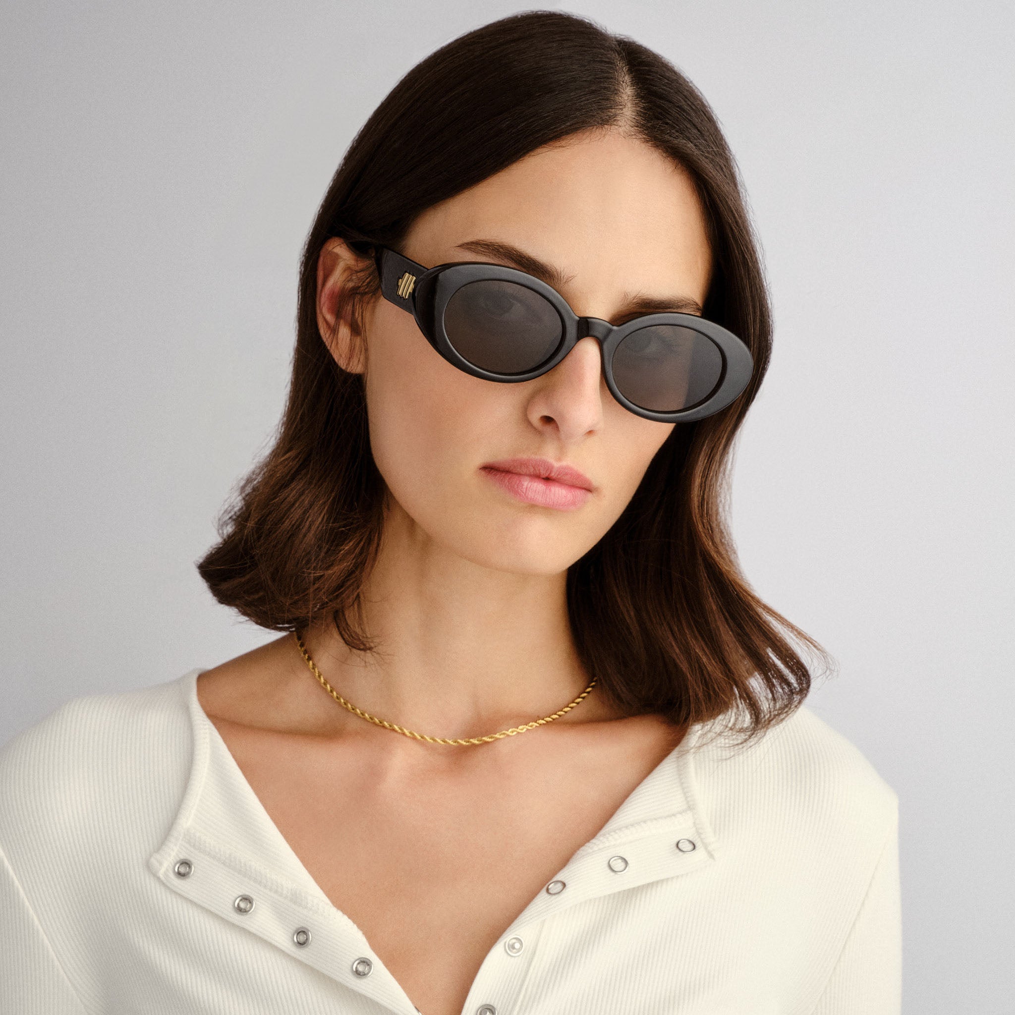 Model wears black oval sunglasses with thick frames and a gold logo paired with a slim fit white ribbed top.