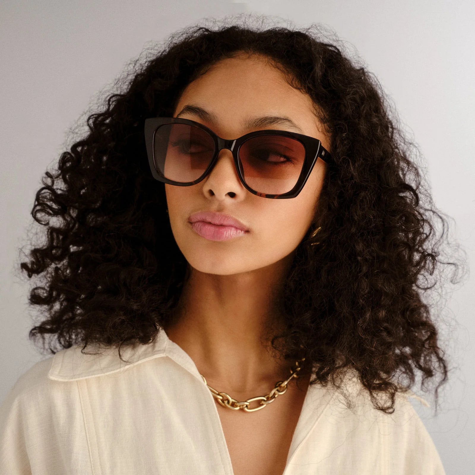 Oversized dark square sunglasses with gradient lenses paired with a relaxed cream collared shirt and gold chain.