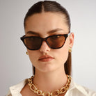 Tortoiseshell cat eye sunglasses feature brown lenses and a sleek fit worn with a chunky gold chain and white shirt.