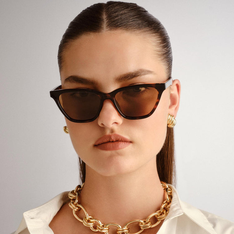 Tortoiseshell cat eye sunglasses feature brown lenses and a sleek fit worn with a chunky gold chain and white shirt.