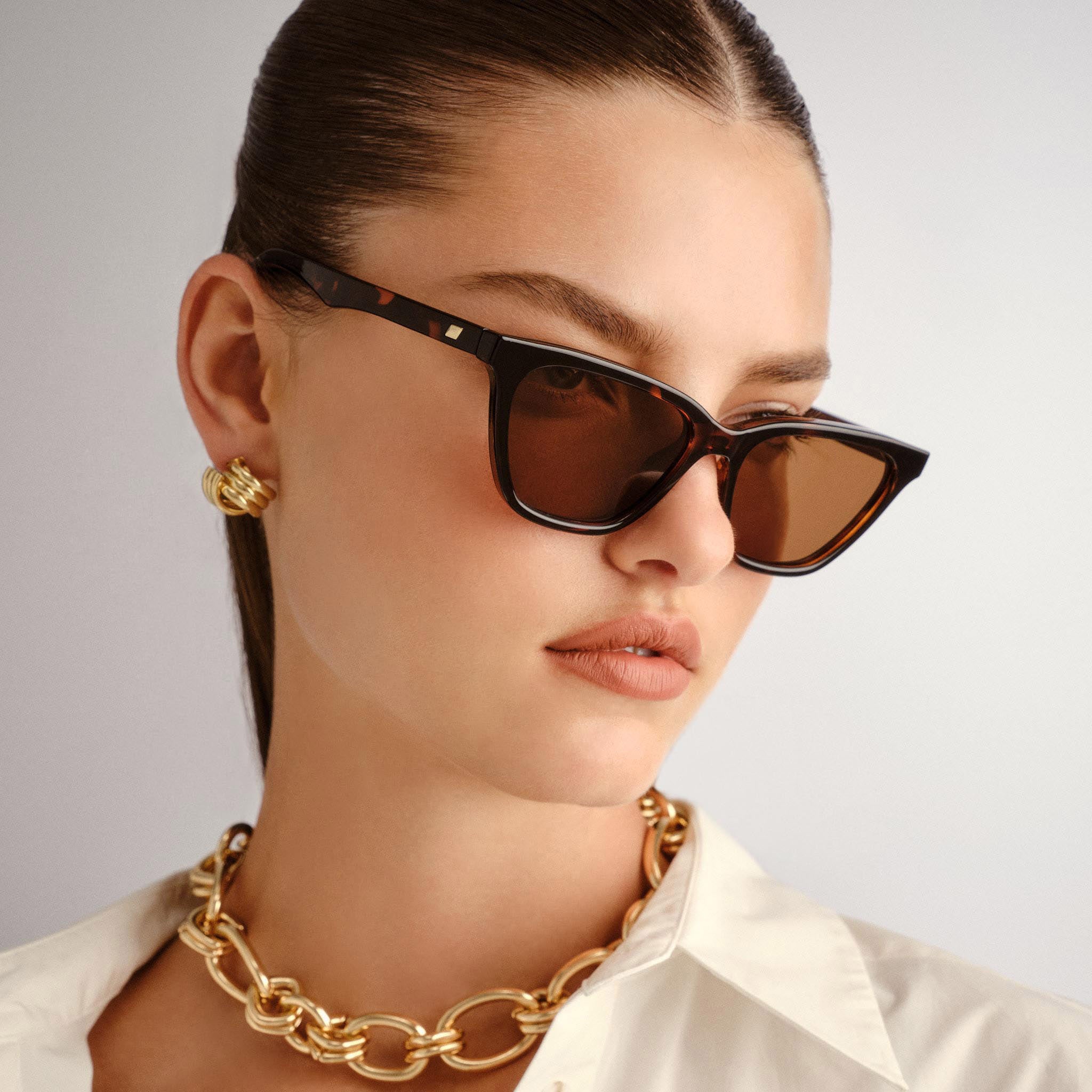 Model wears angular tortoiseshell cat eye sunglasses with brown lenses a chunky gold necklace and a white shirt.