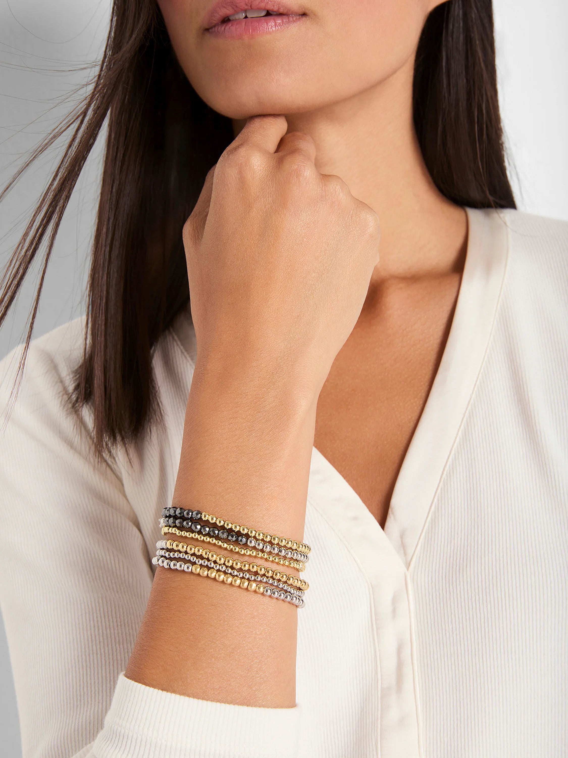 Stacked gold, silver, and dark gray beaded bracelets with varied textures, worn on a model's wrist.
