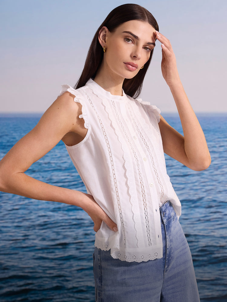 Woman wearing a white sleeveless top with ruffled details, standing by the ocean.