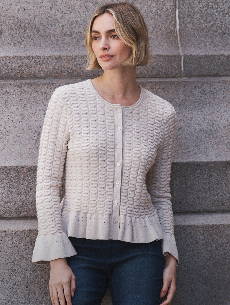 A woman with a short blonde bob wearing a textured cream cable-knit cardigan with button-front closure and flared cuffs, paired with dark blue jeans, standing against gray stone steps.