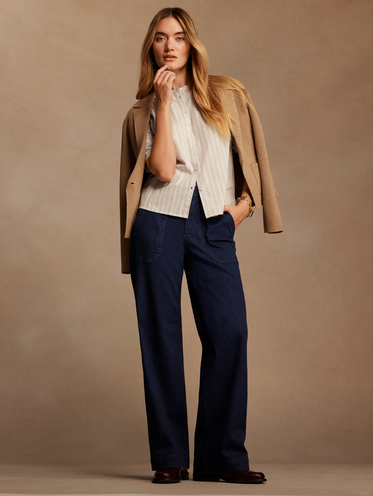 A woman models a stylish outfit consisting of a cream ribbed knit top, a tan blazer, and dark navy wide-leg trousers, set against a solid warm beige studio backdrop.