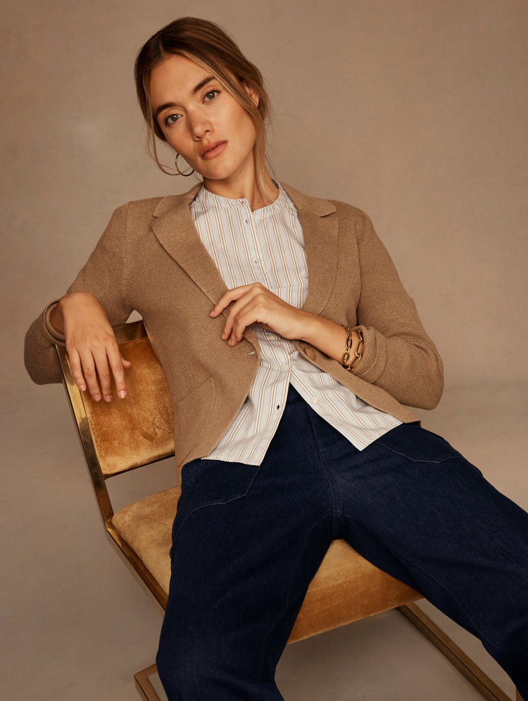 A woman models a tan wool-blend blazer over a white striped button-down shirt and dark blue jeans, seated on a modern wooden chair against a warm beige studio backdrop.