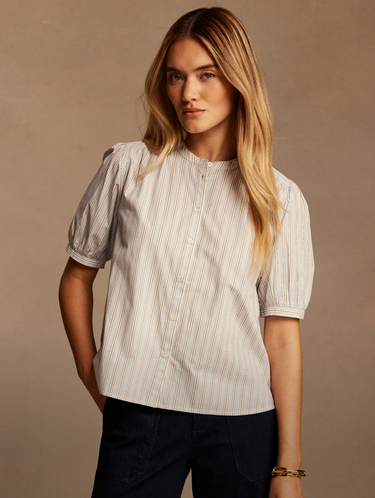 A woman modeling a short-sleeve button-front blouse with vertical pinstripes, photographed against a plain warm beige studio background.
