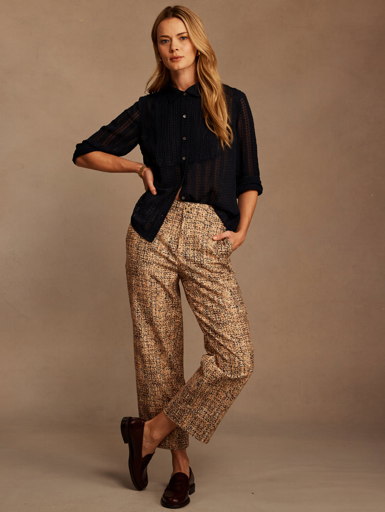 A woman modeling a dark navy blue button-up shirt and beige-and-brown patterned trousers, standing against a solid taupe studio backdrop.