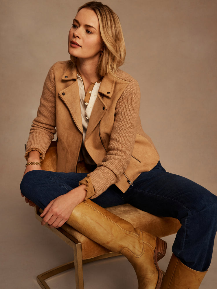A woman sits on a tan upholstered chair wearing a camel-colored suede moto jacket over a white top, dark blue jeans, and tan leather boots, against a warm beige studio backdrop.