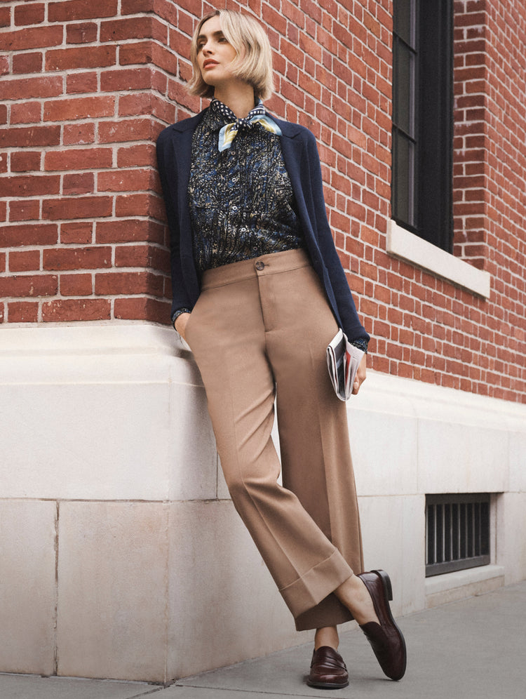 A woman stands outdoors against a red brick building wall, wearing a navy blazer, a dark patterned blouse with a light scarf, tan wide-leg cropped trousers, and dark brown loafers, while holding a small clutch.