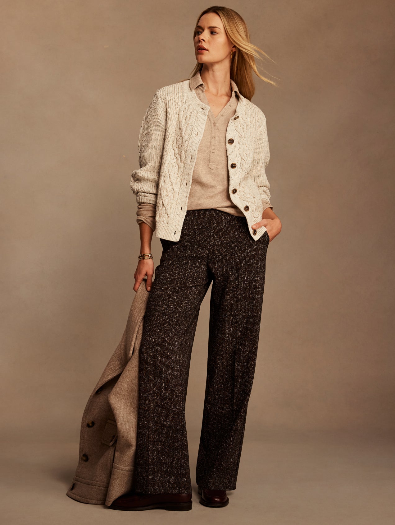 A woman models a stylish outfit featuring a cream cable-knit cardigan over a beige top, paired with dark brown wide-leg tweed trousers and a draped beige coat, set against a warm beige studio backdrop.