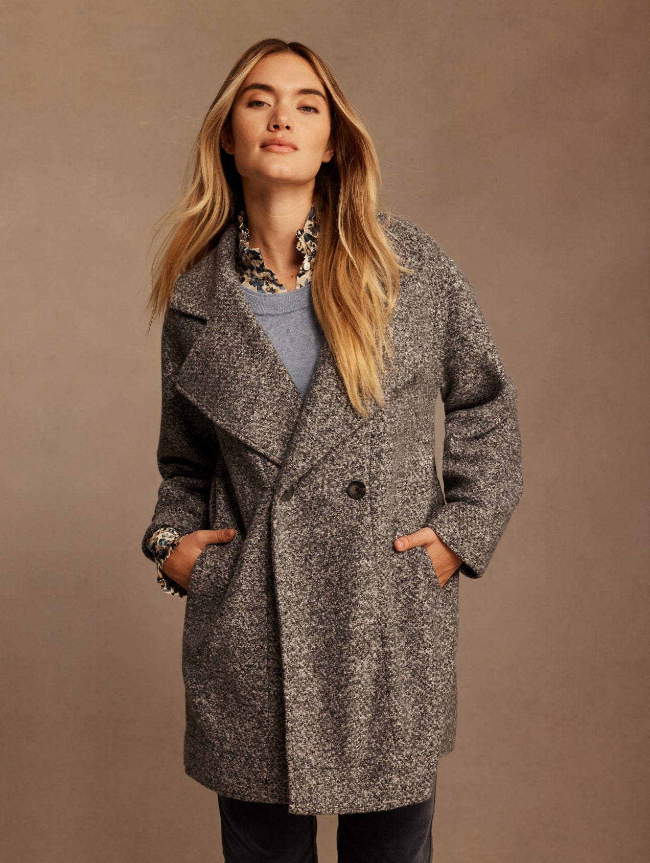 A woman modeling a textured gray and black tweed double-breasted coat over a light blue top, set against a solid warm brown studio background.