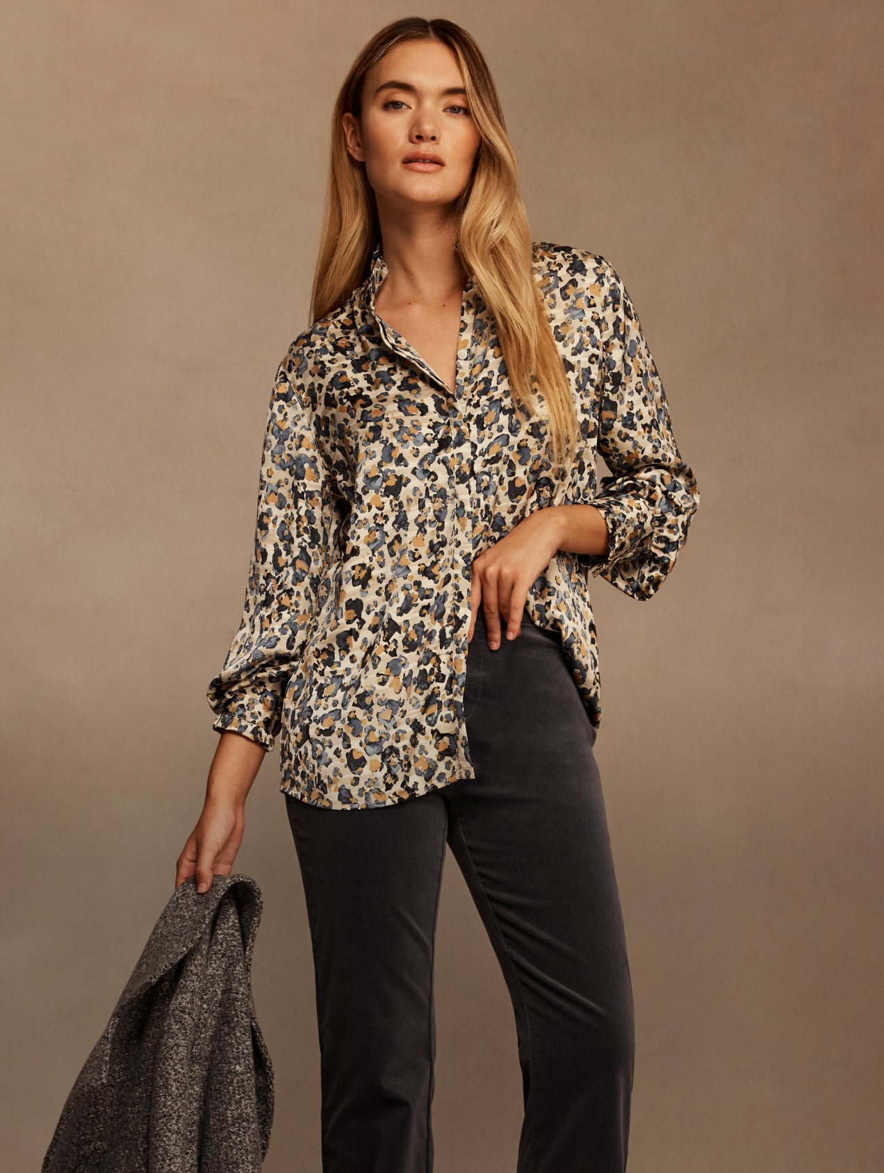 A woman modeling a long-sleeve button-up shirt with a beige, black, and blue leopard print, paired with dark charcoal trousers, holding a gray jacket against a warm brown studio backdrop.