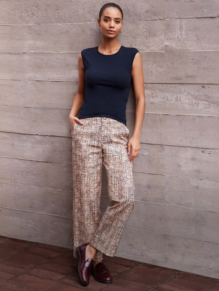 A woman stands against a gray concrete wall wearing a sleeveless navy top and patterned beige wide-leg trousers with dark brown loafers.