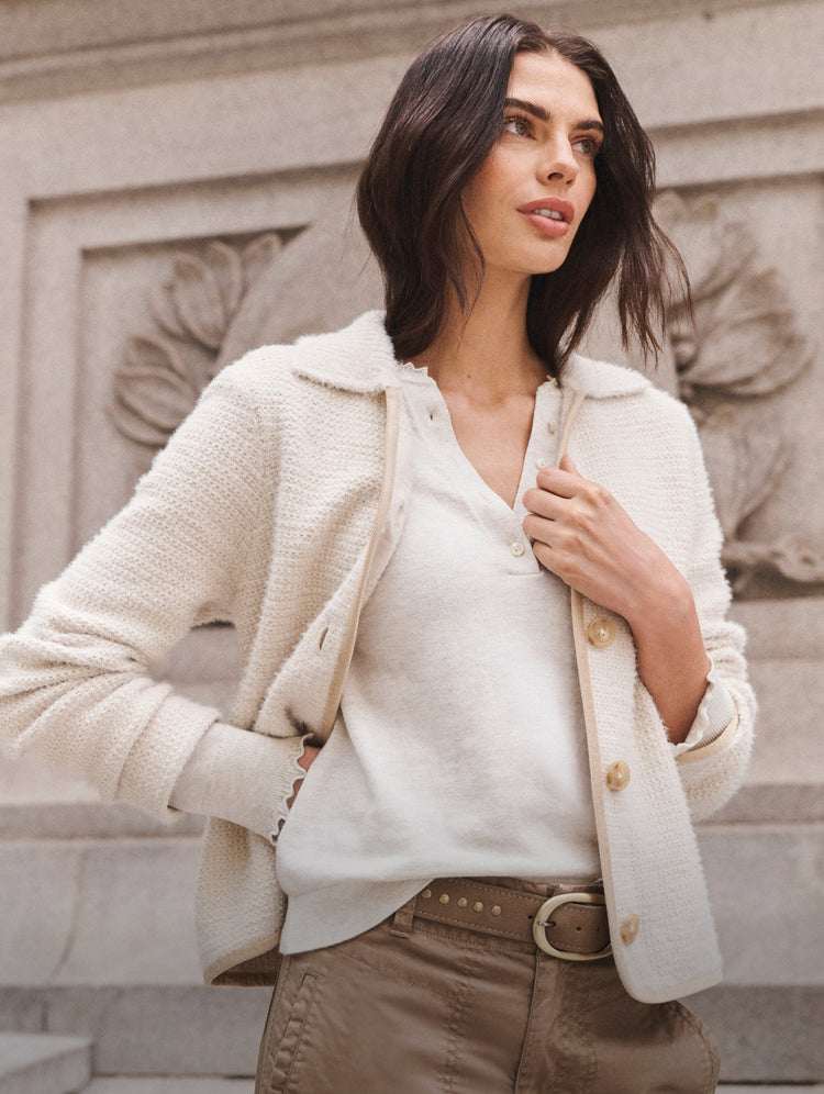 A woman wearing a cream-colored textured knit cardigan over a matching cream V-neck top, paired with brown trousers and a brown belt, standing outdoors against a stone wall.