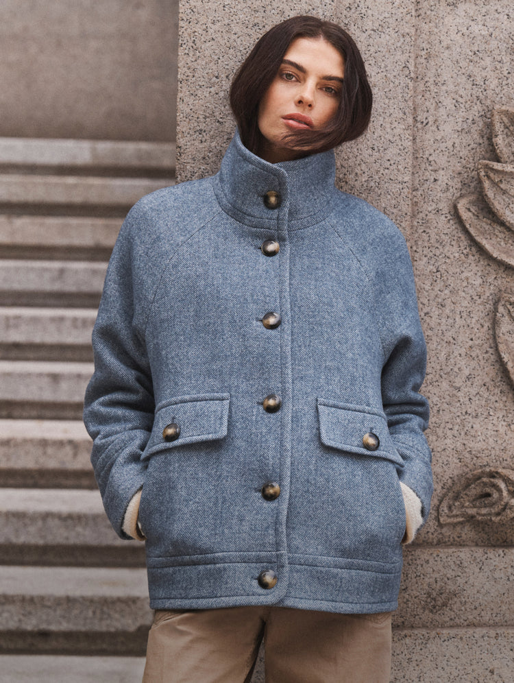 A woman wearing a light blue heathered wool-blend jacket with a high stand collar, large front buttons, and flap pockets, standing outdoors against a stone wall and steps.