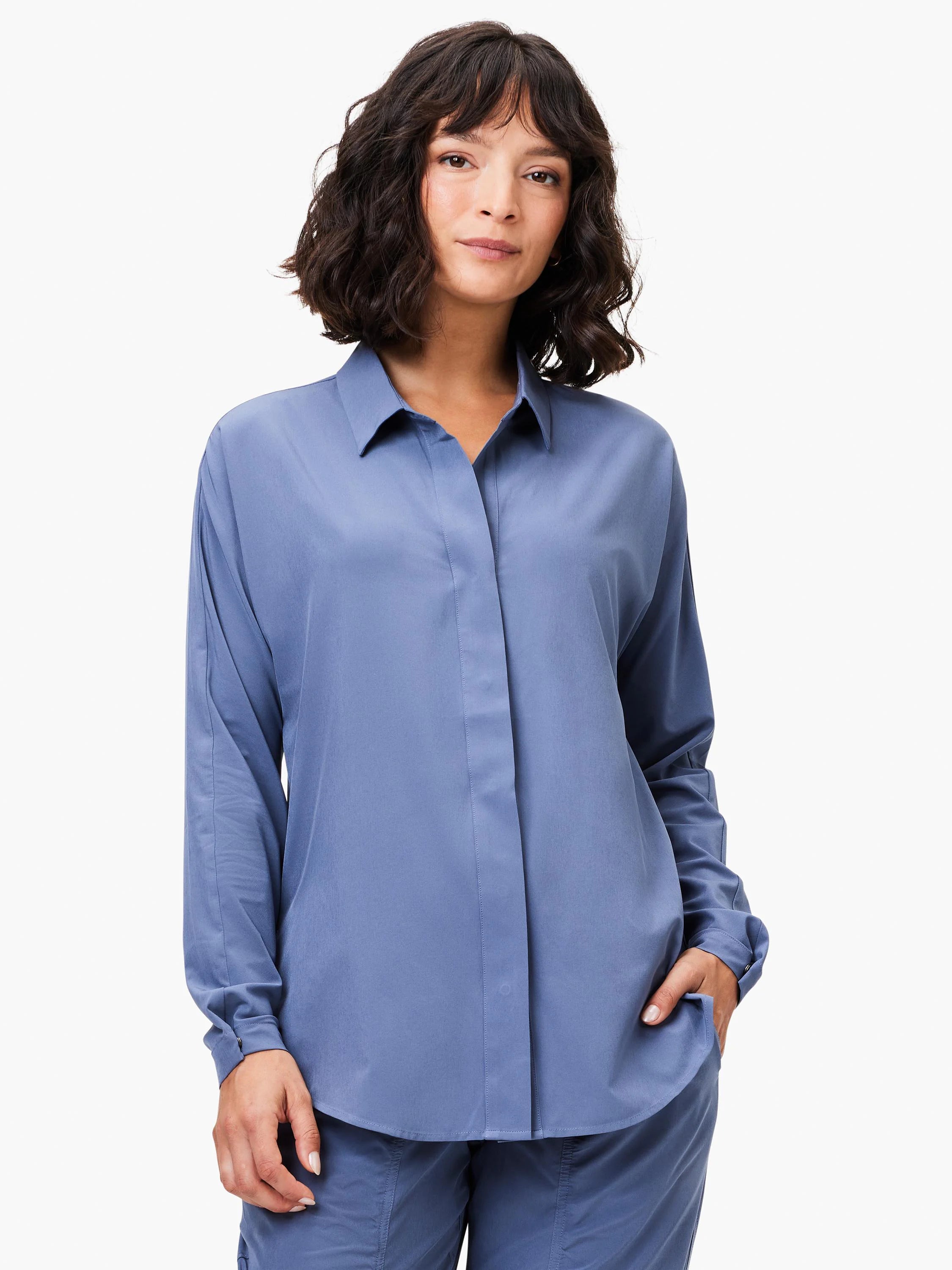 Woman wears a dusty blue collared tech stretch shirt with long sleeves and a relaxed, untucked fit.