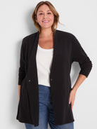 Model wears a relaxed fit black longline knit cardigan with a draped collar and hidden snap closure.