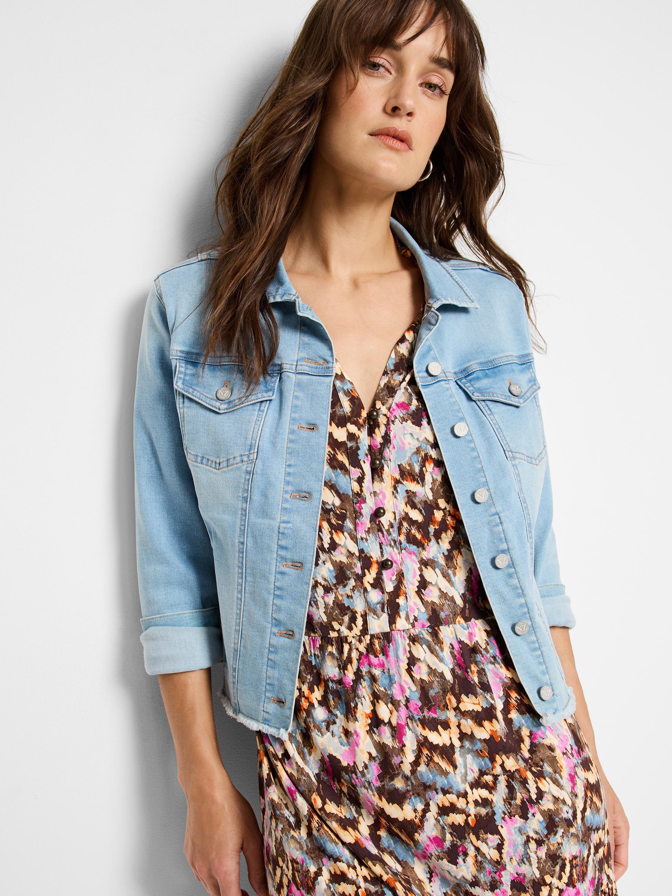 A light wash fitted denim jacket featuring rolled three-quarter sleeves and a subtle raw hem is worn over a printed dress.