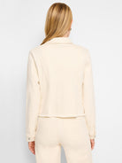 Model viewed from the back wearing a slightly cropped, light beige denim jacket and matching light beige denim pants. The jacket has a traditional collar, long sleeves with button cuffs, vertical seam detailing on the back, and a raw, frayed hemline.