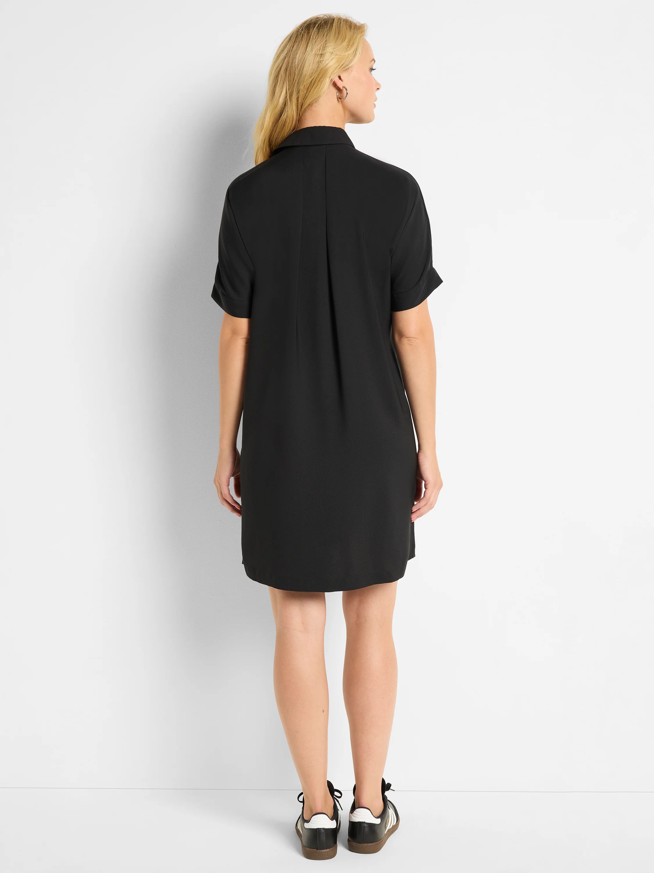 Black shirt dress, back view. Collared, short cuffed sleeves, loose-fitting, above-knee length, with back pleats.