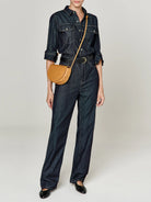 Tan half-moon crossbody bag with a dark denim jumpsuit.  The bag has a light brown leather strap.
