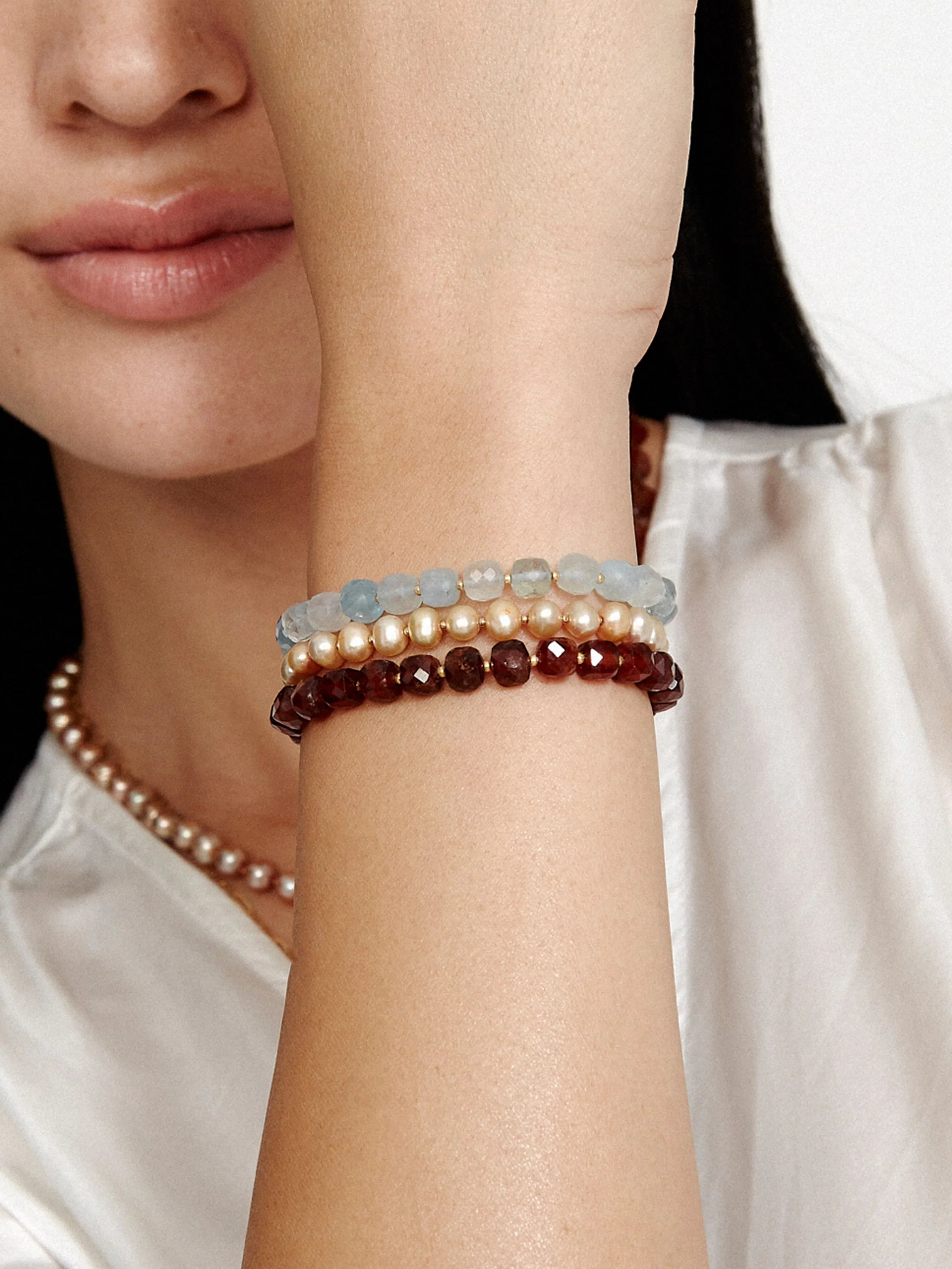 Three stacked bracelets. Top: light blue stones. Middle: beige pearls. Bottom: dark reddish-brown stones. Gold accents.