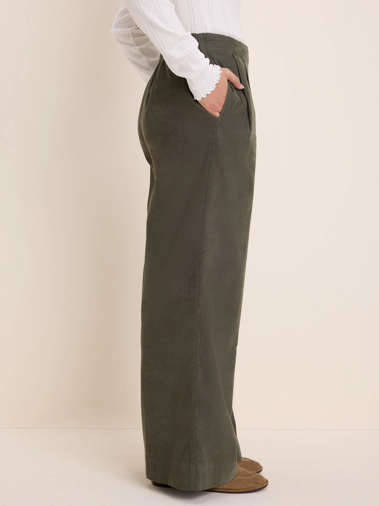 Dark olive green baby corduroy wide-leg pants with pleated waist. Model's hand in one front pocket.