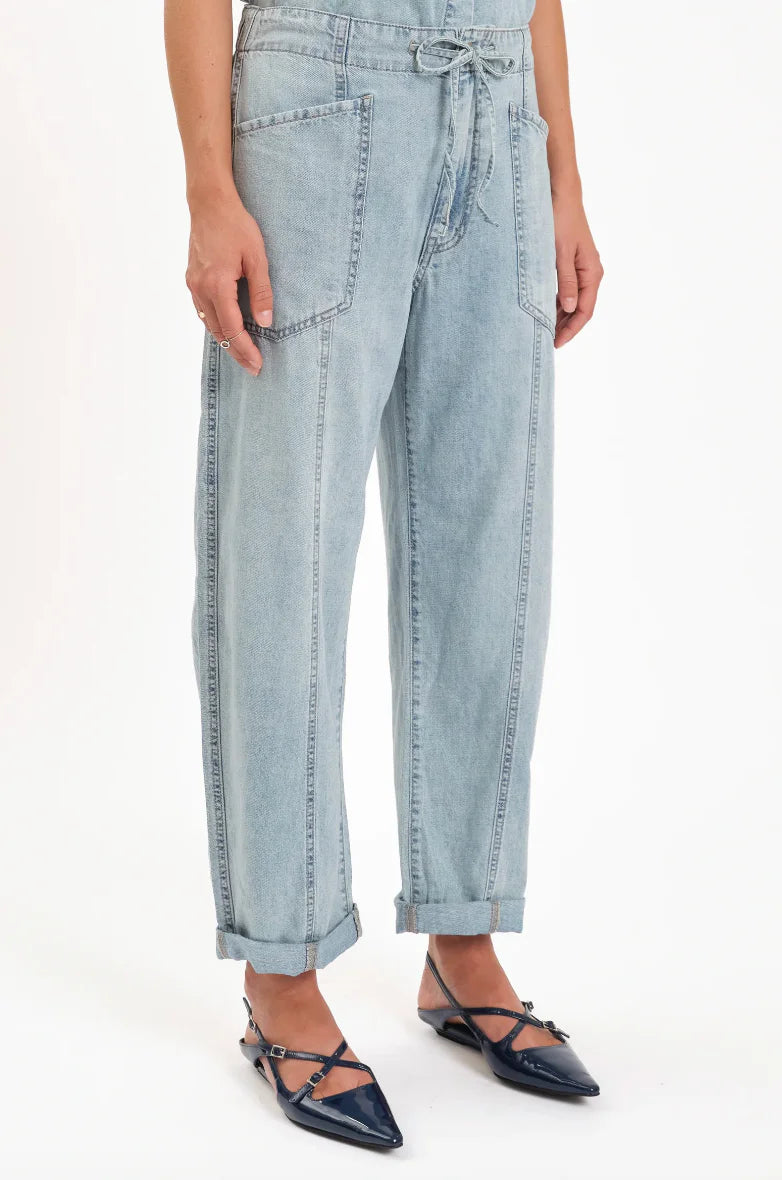 Light wash blue barrel leg jeans with tied drawstring, large front patch pockets, exposed vertical seams, shown cuffed.