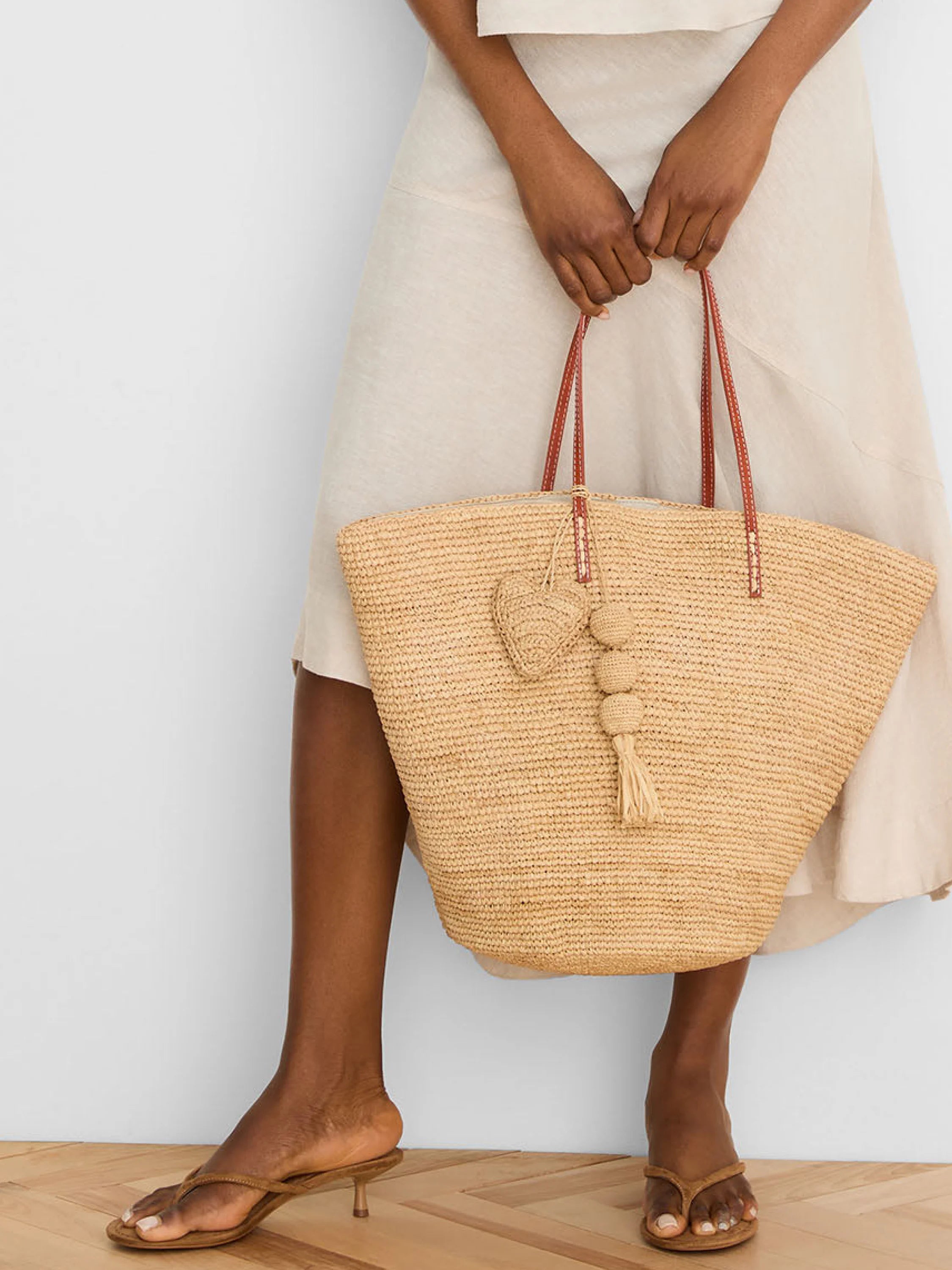 Beige woven tote bag with dark brown straps. Features a matching woven heart charm & a beaded tassel charm.