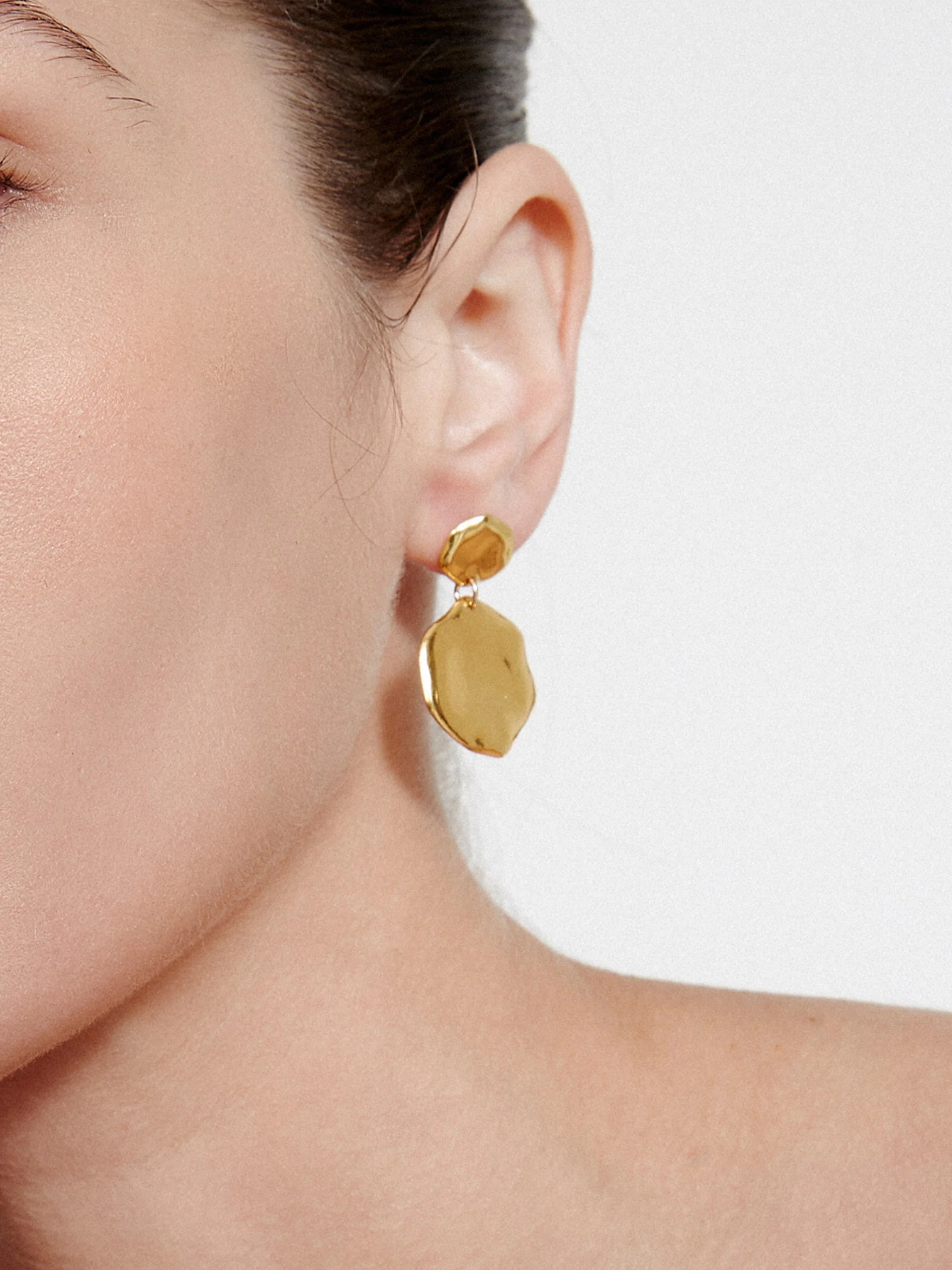 Gold-toned hammered drop earring with two irregular, textured metal pieces, worn on an ear.