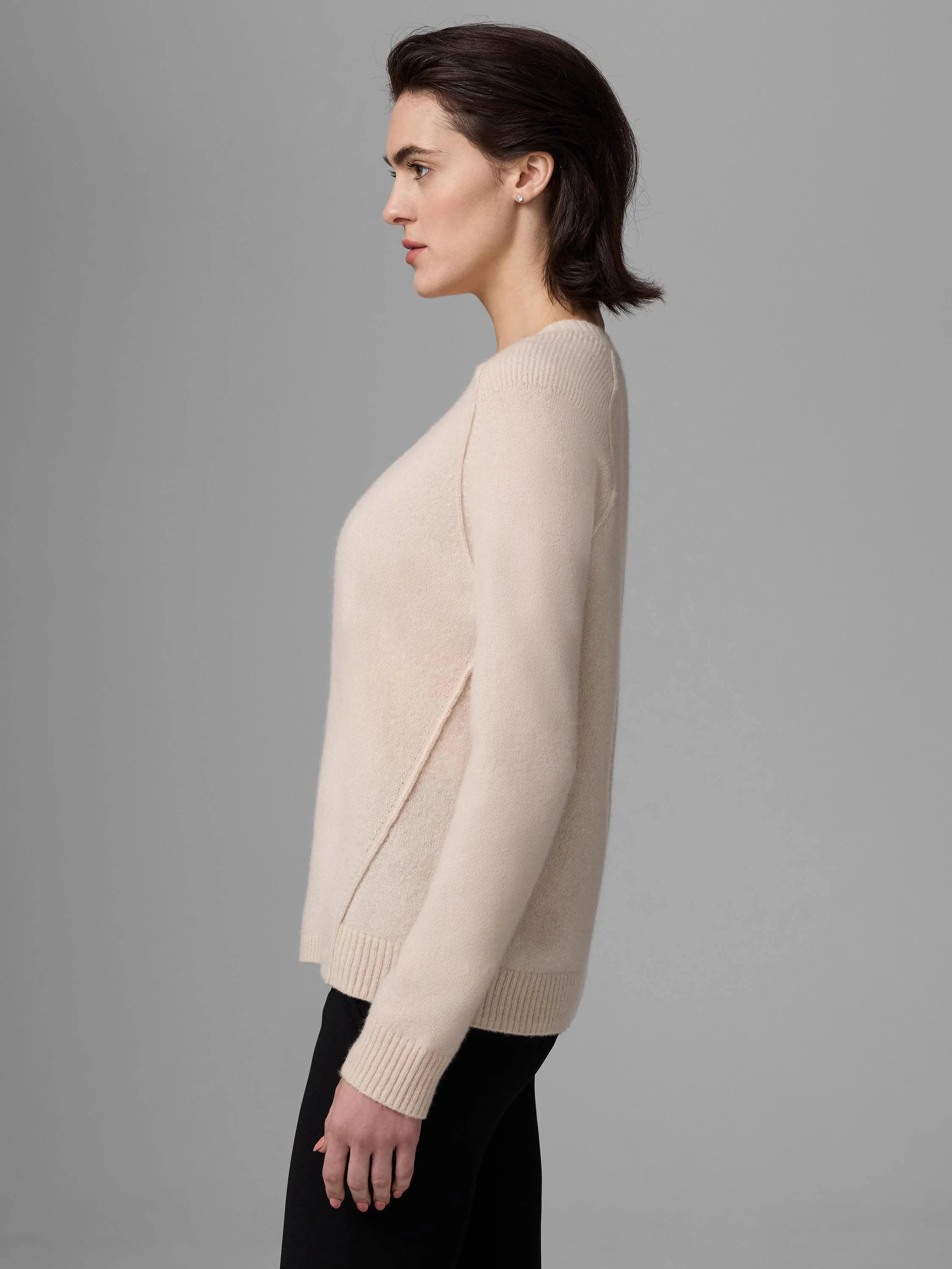 Soft, light beige sweater with ribbed cuffs, hem, and angled side slits. Model in profile.