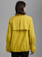 Back of a woman wearing a yellow twill trench coat with a pleated storm flap, buckled cuffs, and an elasticated hem.