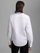White long-sleeve shirt, back view on model. Features buttoned cuffs and a curved hemline.