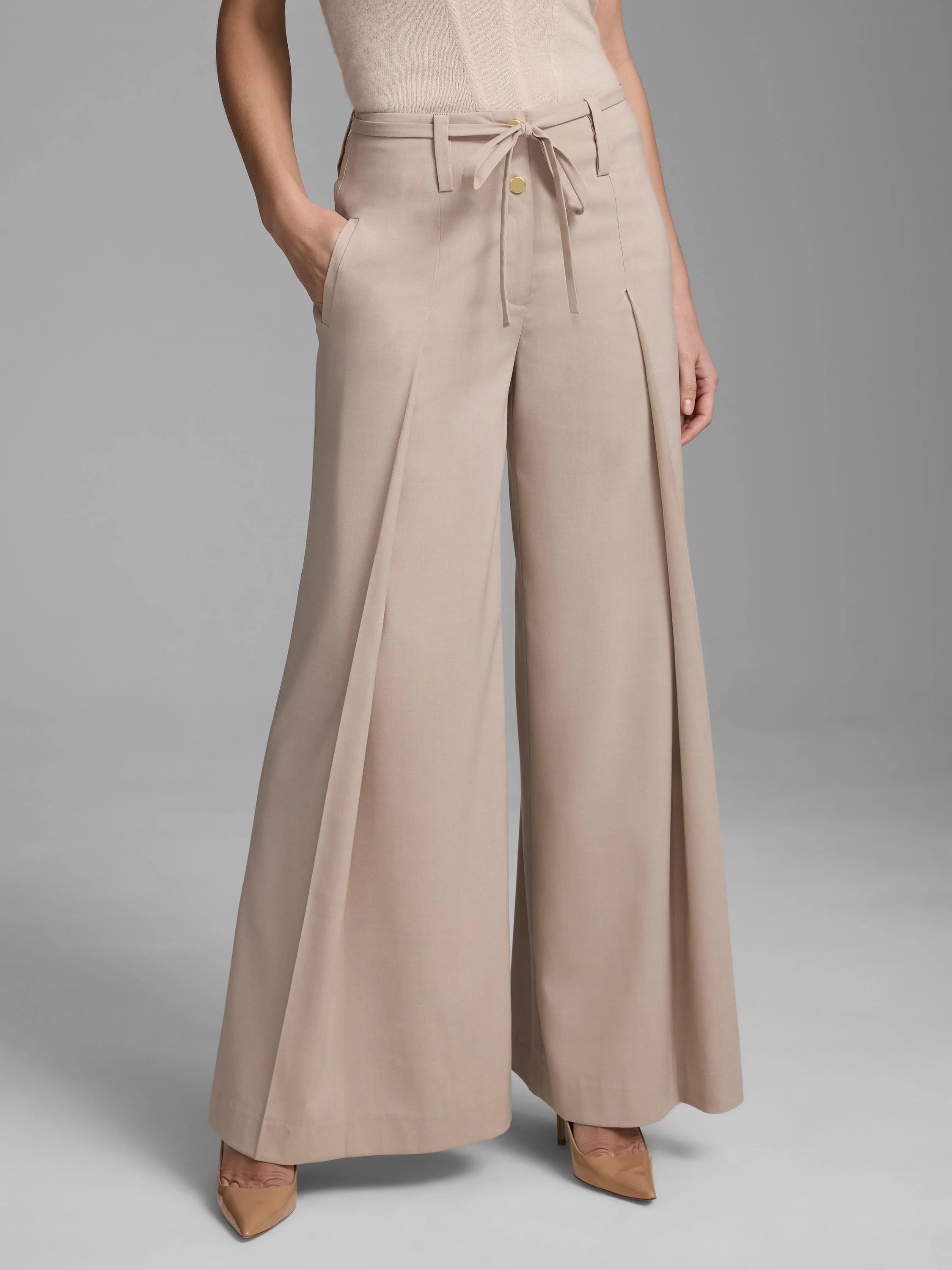 Light tan wide-leg pants with deep front pleats, a fabric tie-belt, and a gold button closure at the waist.