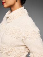 Cream jacket with a textured base, featuring distinct rows of fringe on the sleeves and body. A silver zipper pull is...