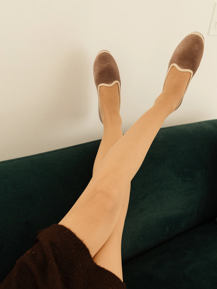 Brown velvet slippers with a cream-colored trim, shown on a pair of legs in beige fishnet stockings.