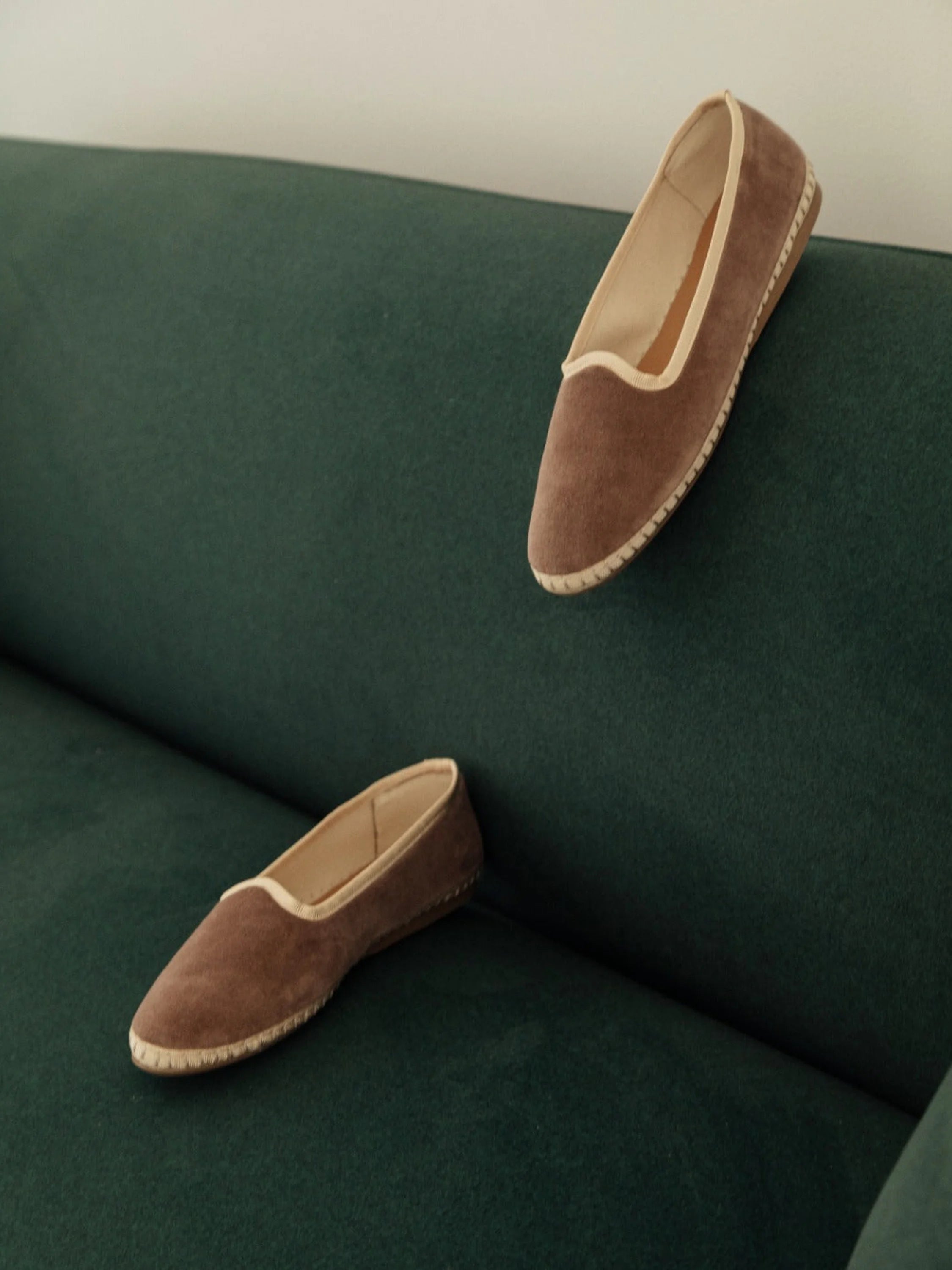 Two taupe velvet slippers with beige trim, resting on a dark teal sofa.