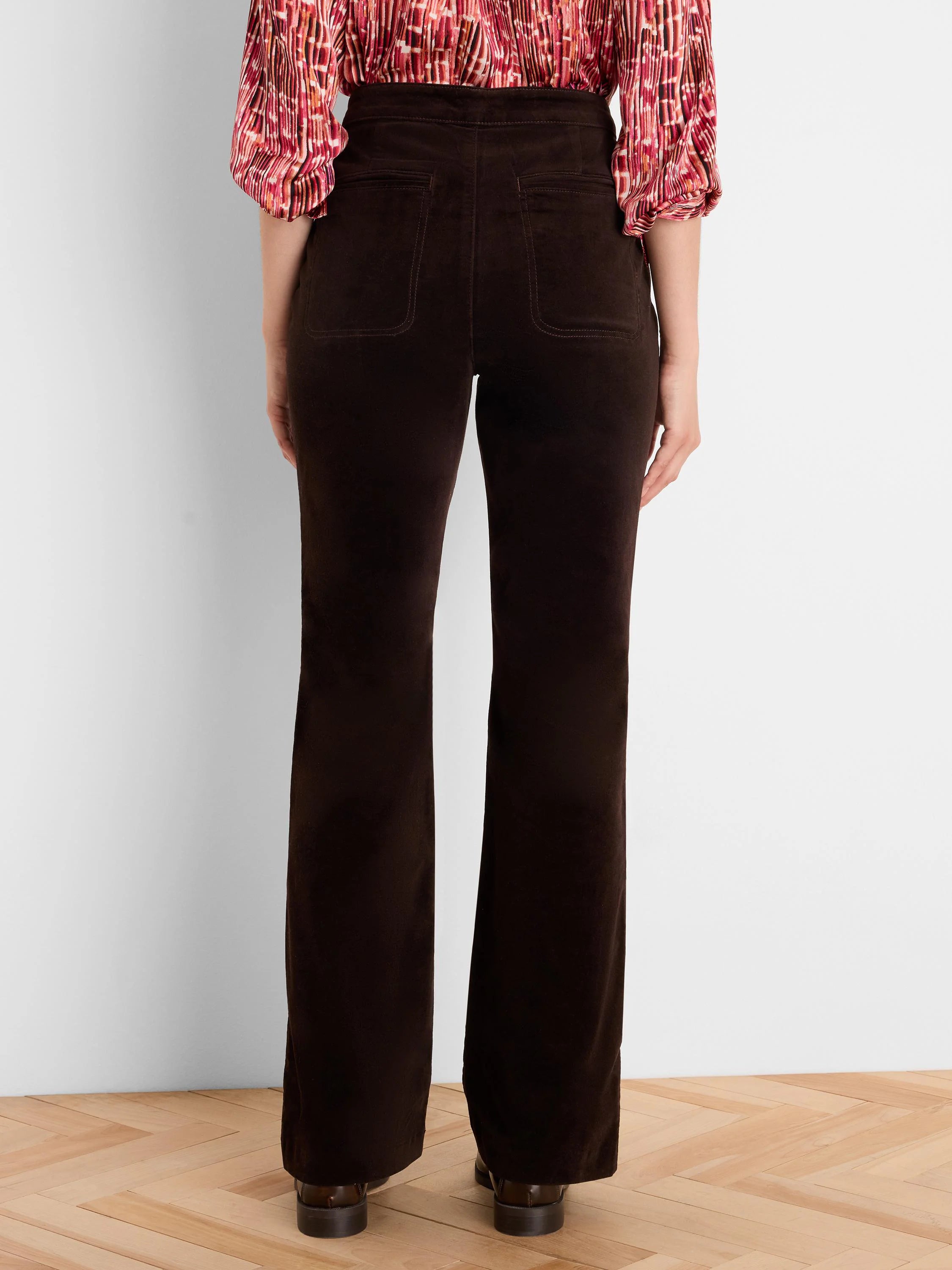 Dark brown velvet boot-cut pants on model from behind. Features back patch pockets and a subtle leg flare.