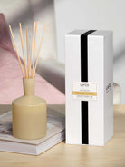 Round pale yellow diffuser bottle with light brown reeds. White product box features a black vertical stripe and yellow...