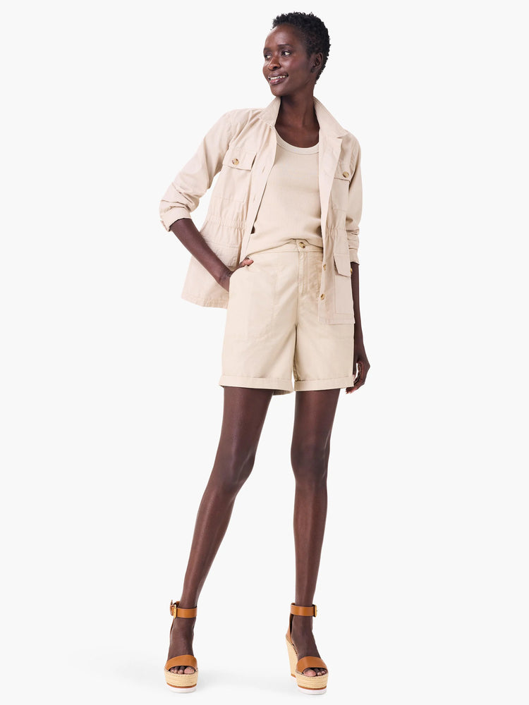 Pale tan poplin jacket with four button-flap pockets, collar, and cinchable waist, worn open over a matching top.