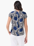 Back view of a short-sleeve top with gathered detail. Features a complex pattern of white petals and blue dots on a dark...