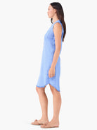 Light blue linen sleeveless shirt dress, asymmetrical hem with frayed edges. Model wears nude sandals.