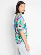 Blue top with vibrant green, orange, and pink abstract leaf print. Short sleeves, slightly puffed.