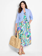 Model wears light blue cardigan over midi dress with vibrant tropical print in pinks, oranges, greens, and blues.  She...